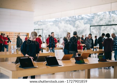 Young female customer standing near table with electronic products while examining the goods, store employees and visitors stand in blur on background. Millennial hipster girl checking device for buy