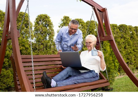 Elderly couple sitting on a swing in the garden and attending a video call via laptop. Elegant older people look at the screen, wave their hands and drink tea from a cup.