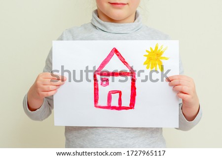 Closeup of Hands of child holding picture of red house with sun covering her face on yellow background. The child learns to draw.