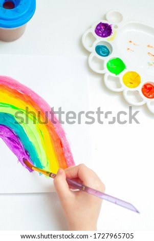 Top view close up of a hand of child drawing a rainbow on the white paper. A kid paints at home.