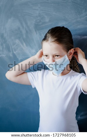 Girl schoolgirl puts on a medical disposable mask, hands are raised against the background of the school blackboard. Vertical image