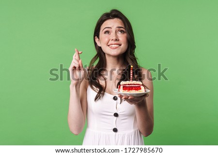Image of excited young woman with fingers crossed holding cake with candle isolated over green background