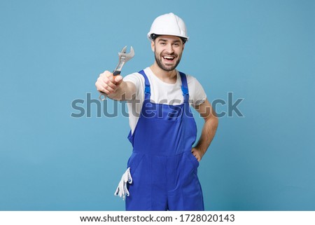Cheerful young man in coveralls protective helmet hardhat hold adjustable wrench isolated on blue wall background in studio. Instruments accessories for renovation apartment room. Repair home concept
