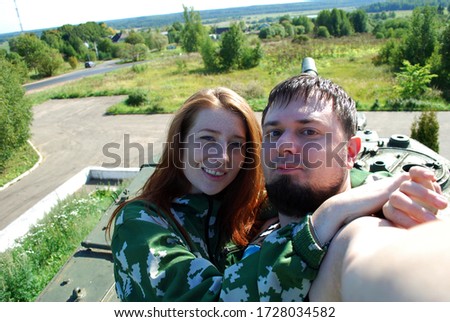 A young couple on a hot summer day takes a selfie against the background of a beautiful landscape behind them while standing on a tank. A young girl and a guy with a beard are photographed.