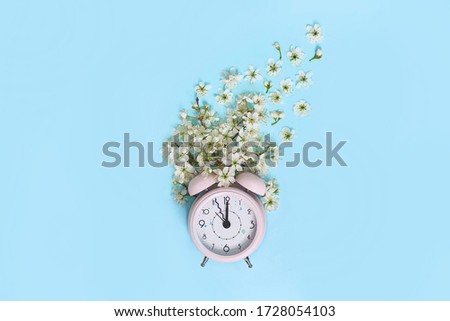 Pink alarm clock and delicate little white flowers on blue background. Top view. Time for love and greetings. Spring Time Change, Spring flowers and Alarm Clock.