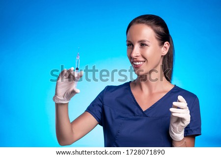 Portrait of a brunette with a braided tail, posing while standing against a blue background, in a blue surgical suitin medical gloves, squeezing a medicine from a syringe.
