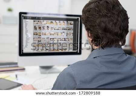 View from behind over his shoulder of a photographer editing photos on a computer with rows of images visible on the monitor
