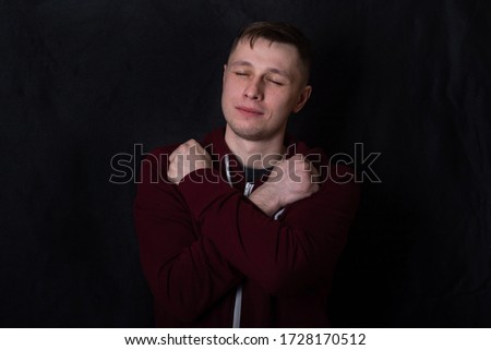 Studio portrait of a young man in a red sweatshirt against black background. A deaf-mute guy shows a "love" gesture with his fingers. Good eyes. Facial expression and sweet smile. Sign language. Royalty-Free Stock Photo #1728170512