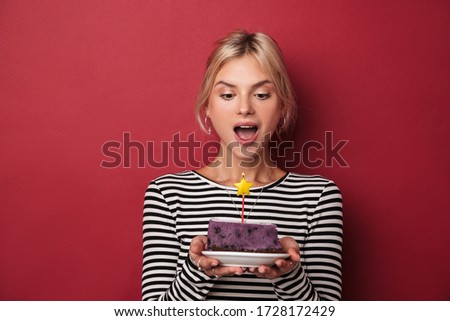 Image of nice delighted woman in striped sweatshirt holding cake with candle isolated over red background