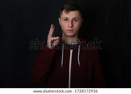Studio portrait of a young man in a red sweatshirt against black background. A deaf-mute guy shows a "water" gesture with his fingers. Good eyes. Facial expression and sweet smile. Sign language. Royalty-Free Stock Photo #1728172936