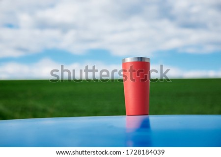 Close-up picture of red thermos cup with tea, standing on blue car top in front of green field and light blue sky with white clouds Picnic in countryside. Nature protection concept.