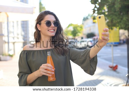 Smiling beautiful brunette woman walking outdoors by street, take selfie by mobile phone, drinking juice.