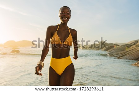 Attractive african woman in yellow bikini walking along the beach. Smiling female in swimwear coming out of the sea water at sunset.