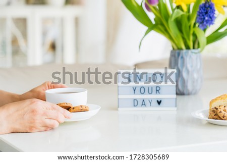 Good morning concept, breakfast time. Close up woman with cup of hot coffee drink, cookies, lightbox with message Enjoy your day and fresh flowers in vase on table with light interior view. Copy space