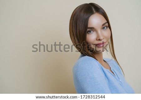 Profile shot of lovely dark haired woman with European appearance, has healthy well cared skin, minimal makeup, wears blue jumper, stands against beige background, copy space for your promotion
