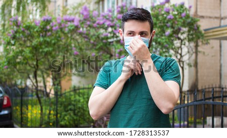 Young handsome man in green t-shirt puts surgical mask on his face, standing outdoor on a background of building and green park. Prevention of viral diseases, coronavirus. 