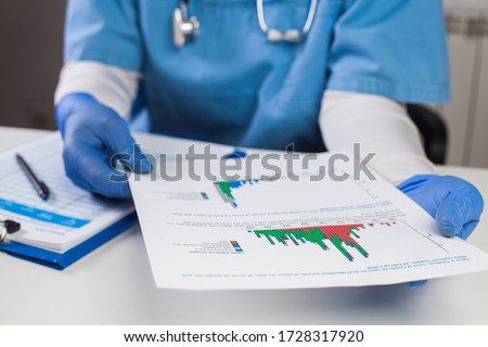 UK doctor wearing protective gloves holding document chart,analyzing COVID-19 graph data,Coronavirus global pandemic outbreak crisis,stats showing number of infected patients,death toll,mortality rate Royalty-Free Stock Photo #1728317920