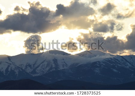 sunset or sunrise with sun glowing behind fluffy blue and white clouds over snowy mountain peaks, blue mountains, and shadowy blue foothills.  Royalty-Free Stock Photo #1728373525