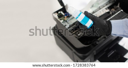 Hand holding cyan color printer ink with inkjet printer in the background. Copy space. Professional printer maintenance concept. Technical assistance in office hardware. Continuous ink system repair.