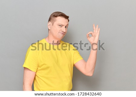 Studio portrait of funny blond mature man wearing yellow T-shirt, with skeptical expression on face, showing okay gesture, expressing his approval and acceptance, standing against gray background