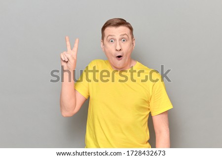 Studio portrait of surprised amazed blond mature man wearing yellow T-shirt, raising hand and showing victory or peace sign, or showing number two with fingers, standing over gray background