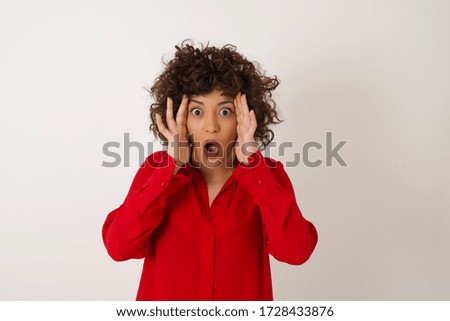 Emotive Arab woman with scared expression, keeps hands on head, jaw dropped, has terrific expression, dressed in stylish red shirt, stands against white background. Omg concept