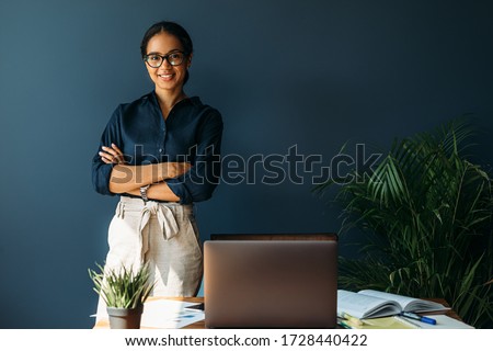 Smiling woman with arms crossed stands at her workplace in home office Royalty-Free Stock Photo #1728440422
