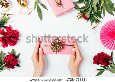 Woman's hands holding pink gift box with golden bow among lush peony flowers and holiday decorations on white background.