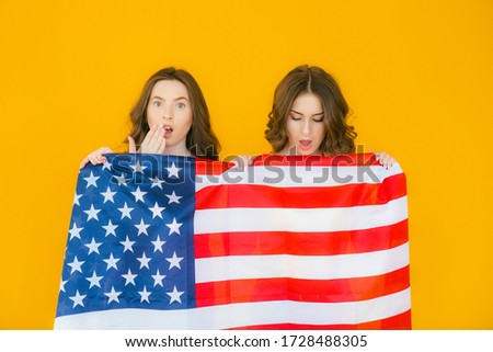 American girls. Two playful young women gesturing peace sign and holding American flag while standing on yellow background