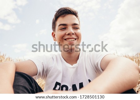 Portrait of cheerful handsome teenage boy, sitting on wheat field, under a sky with clouds, at summer day. Low angle shot. Royalty-Free Stock Photo #1728501130
