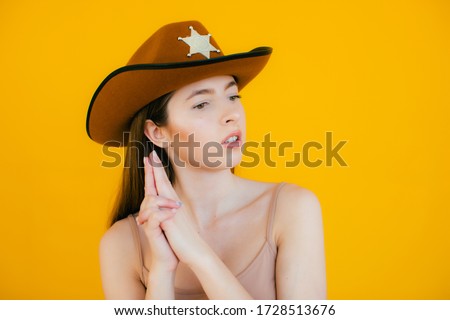 Killing wink. Beautiful young cowgirl gesturing and winking at camera while standing against ton yellow background