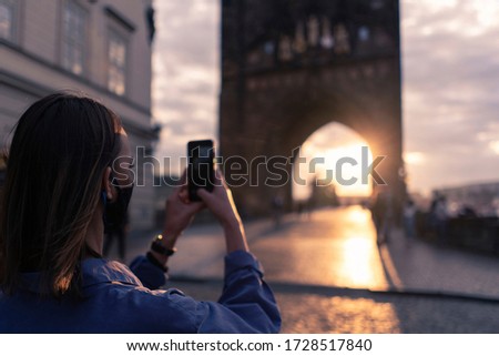 Young woman traveler with medical face mask taking pictures of the Prague, Czech Republic at sunset. Travelling, tourism during coronavirus. COVID-19.
