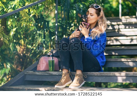 Outdoors lifestyle portrait of pretty girl sitting on the wooden stair in the tropical garden. Hugging and kissing small cute dog. Wearing stylish  jeans, pantone jacket. Relaxing, enjoying life 