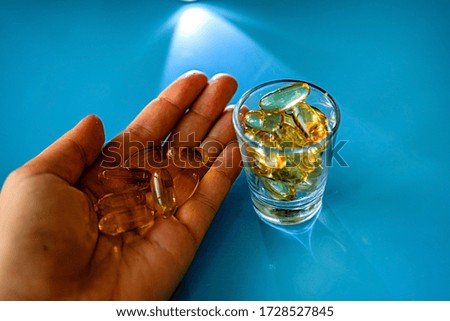 female hand holds a transparent omega 3 capsule, a glass with pills on a blue table
