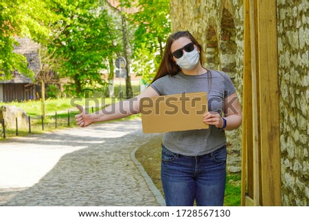 A girl in medical mask with empty cardboard sign stands by the road, catching car for Humorous hitchhiking trip. Getting out of quarantine, traveling after coronavirus. Pandemic fear.