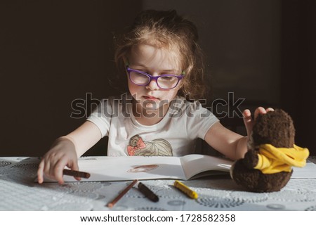 A little girl with glasses draws a portrait of a soft teddy bear, which lies in front of her. Idea for quarantining a child