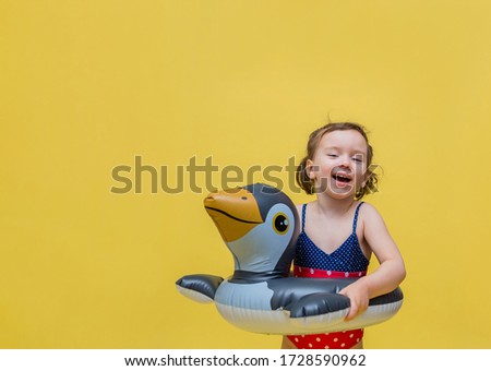 A little girl is laughing with an inflatable circle and in a swimsuit on a yellow background with free space. Inflatable circle in the form of a penguin.