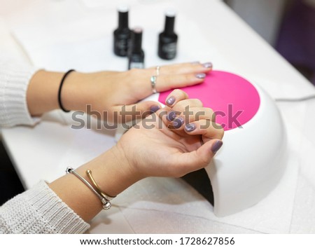 Ready manicure in purple. Hands of a young girl with a gentle purple manicure on a machine for ultraviolet drying gel varnish  in the salon. Nail care after quarantine.