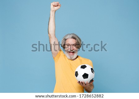 Happy elderly gray-haired mustache bearded man in yellow t-shirt isolated on blue wall background. Sport family leisure concept. Cheer up support favorite team with soccer ball, doing winner gesture