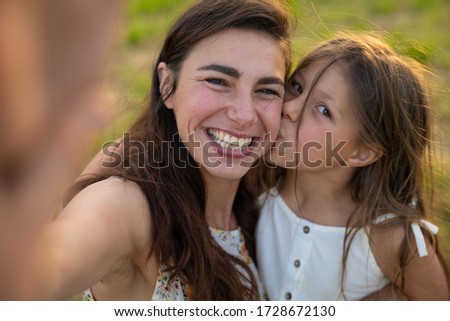 Authentic shot of happy carefree young mother and her little daughter making a selfie or video call to father or relatives on a green nature background in a sunny day.