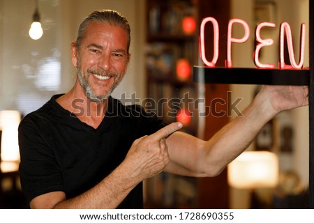 open for business concept, small business owner reopening premises after covid-19 virus pandemic, happy man pointing to red neon open sign at a bar restaurant or cafe window after coronovirus lockdown
