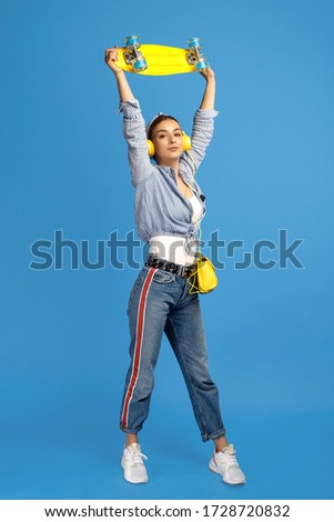 Positive young woman hold over head yellow penny or skateboard over blue background.