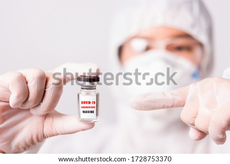Asian female woman doctor or nurse in PPE uniform and gloves wearing face mask protective in laboratory point medicine vial coronavirus vaccine bottle and on bottle has "COVID-19 VACCINE" text label
