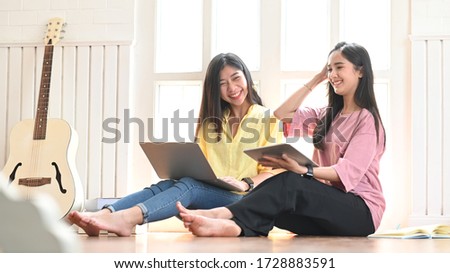 Photo of attractive sisters sitting together on the music studio wooden floor next to acoustic guitar and holding/using a computer laptop and tablet over studio windows as background.