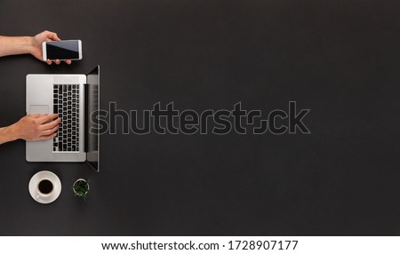Male hands typing on laptop and using smartphone at the same time on abstract dark black office desk with cup of coffe and huge copy space Royalty-Free Stock Photo #1728907177