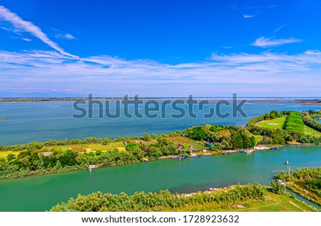 Aerial view of Torcello islands, water canal with fishing boats and green trees. Panoramic view of Venetian Lagoon from bell tower. Veneto Region, Northern Italy. Blue cloudy sky background.