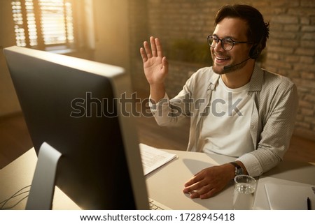 Young happy businessman waving while making video call over desktop PC in the office.