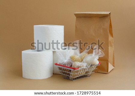 Paper bag, toilet paper and various cereals in small plastic bags in a grocery basket on a brown background. Rice and oatmeal, buckwheat and millet