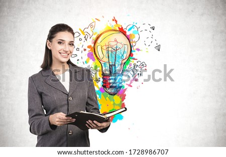 Cheerful young European businesswoman with notebook standing near concrete wall with bright business idea sketch drawn on it. Concept of innovation