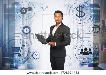Serious young Asian businessman holding laptop in blurry city office with double exposure of hud online shopping interface. Toned image
